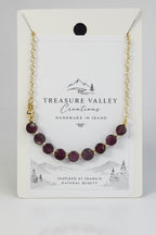 A gold chain bracelet with faceted dark purple beads, presented on a white backing card from Treasure Valley Creations.