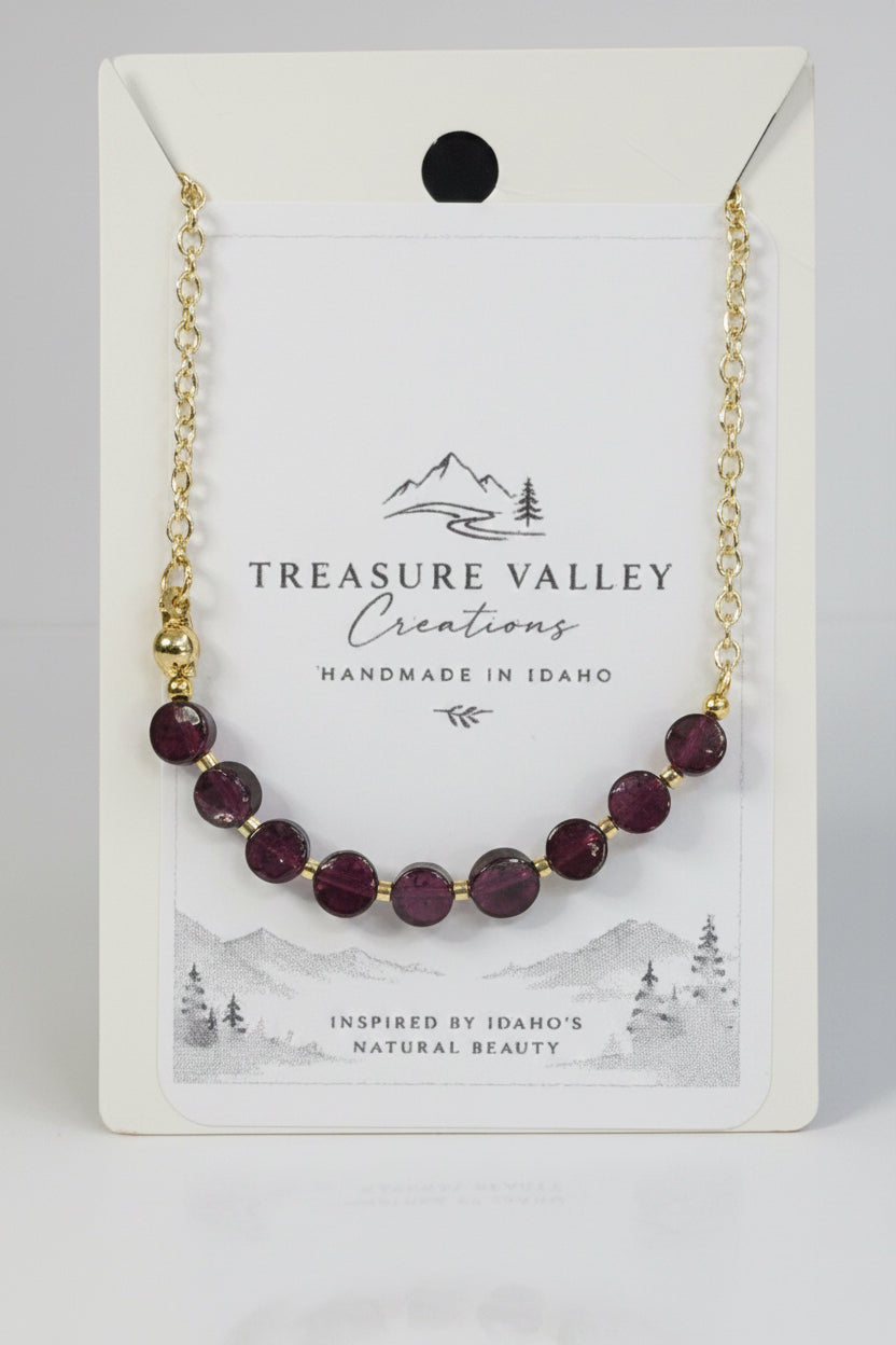 A gold chain bracelet with faceted dark purple beads, presented on a white backing card from Treasure Valley Creations.