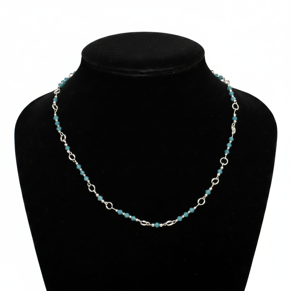 A silver link chain necklace with small turquoise-blue beads, displayed on a black velvet jewelry bust against a dark gray background.