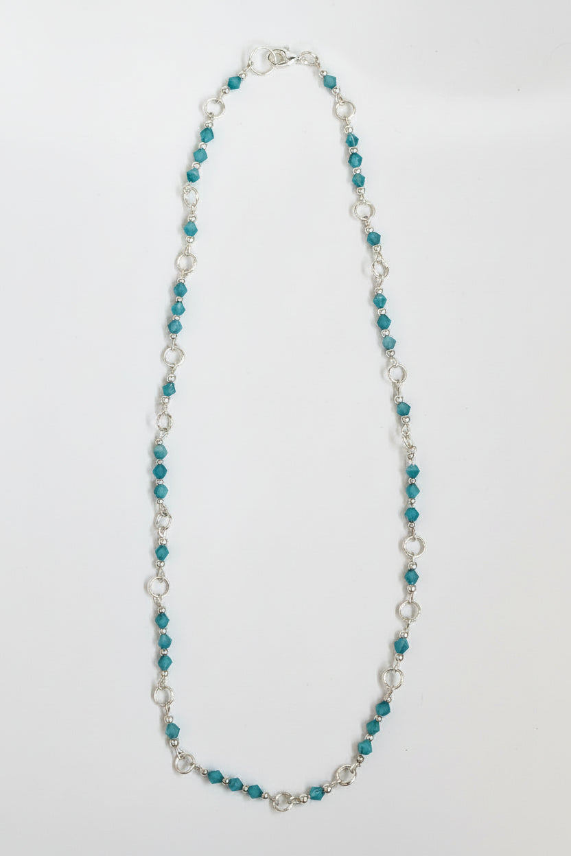 A beaded necklace featuring small teal-blue beads separated by silver-toned rings and a few silver-toned beads, with a silver clasp, shown on a plain light background.