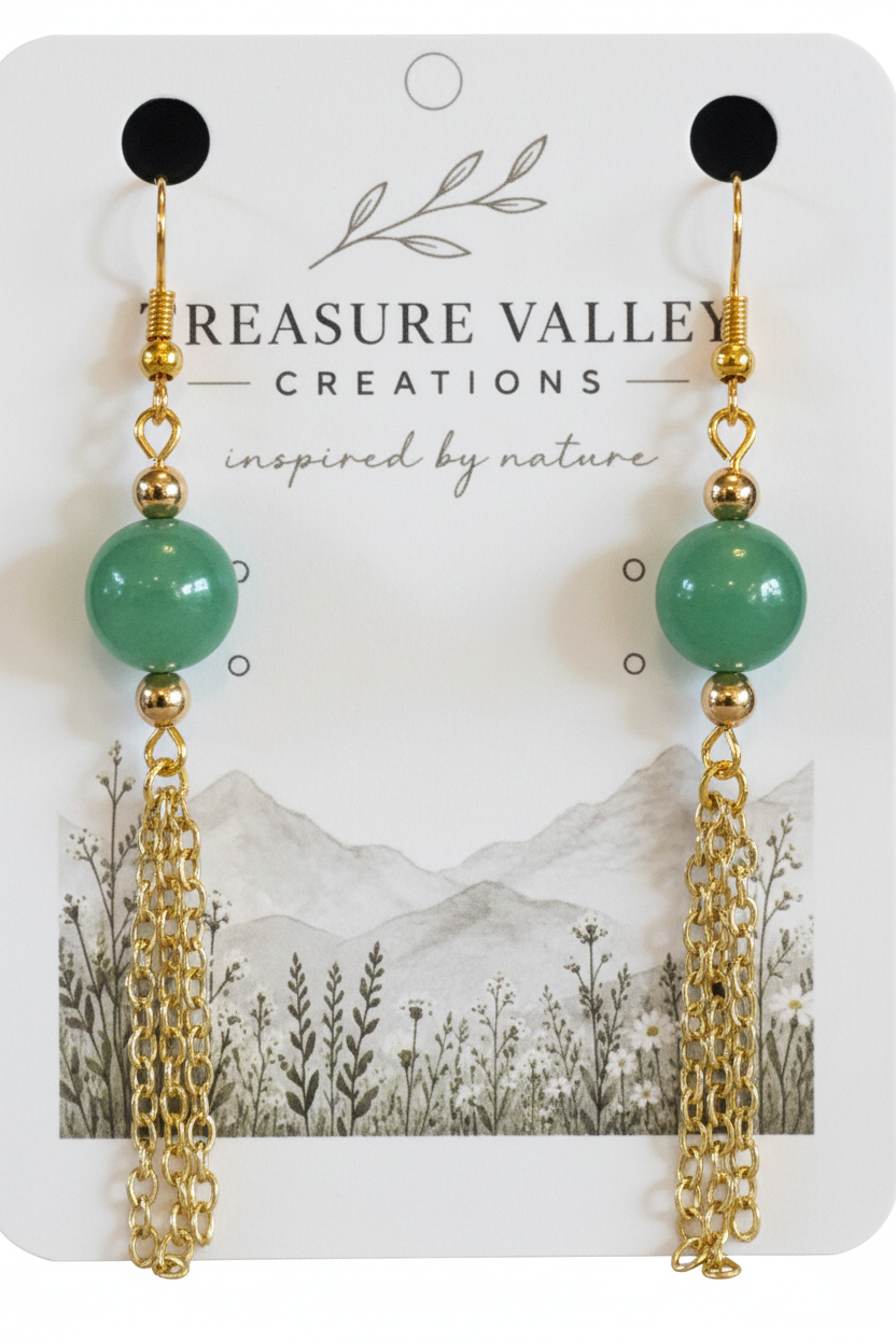 A pair of dangling earrings displayed on a branded card. Each earring has a round mint green bead, gold-toned chain links, and small gold spacer beads.