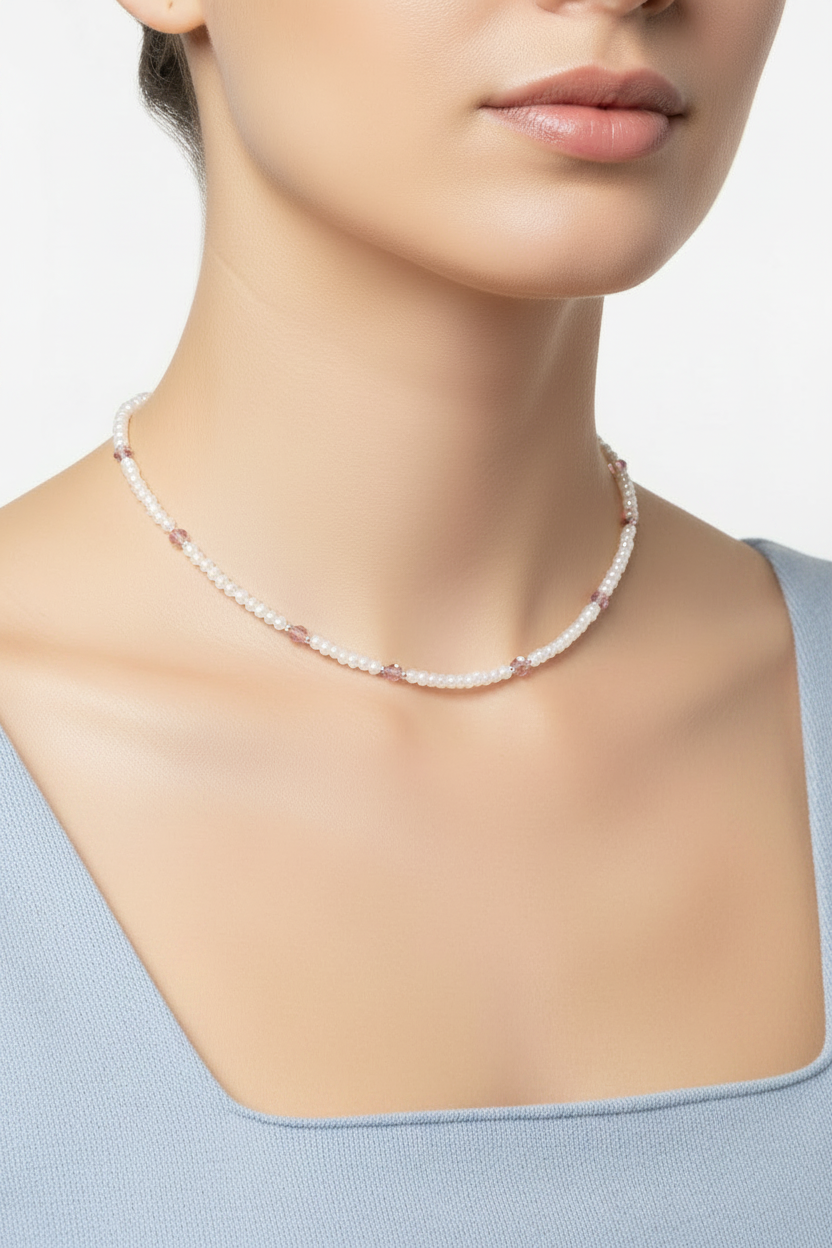 A close-up of a model wearing a delicate necklace made of small white and pink faceted beads, paired with a light blue top against a clean white background.