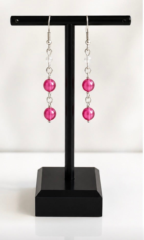 A pair of dangling earrings with bright pink round beads and silver-toned links, displayed on a black T-stand against a white background.