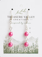 A pair of dangling earrings displayed on a branded card. Each earring has silver-tone hooks, small chain links, and a glossy pink pearl bead near the bottom.
