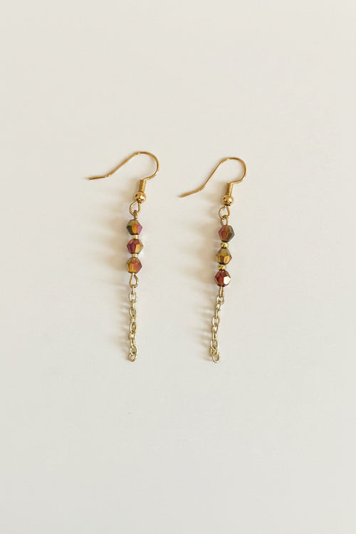 A pair of gold dangle earrings with faceted beads in amber, gold, and pink, hanging from gold fishhook wires on a plain light gray background.