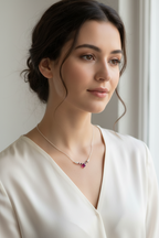 A woman with dark hair styled in an updo wears a delicate silver necklace with a small cluster of pink gemstones and a central teardrop-shaped red stone, paired with a cream satin V-neck top.
