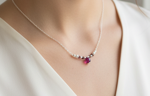 A delicate silver chain necklace worn by a model, featuring a small cluster of clear stones and a single faceted purple gemstone pendant.