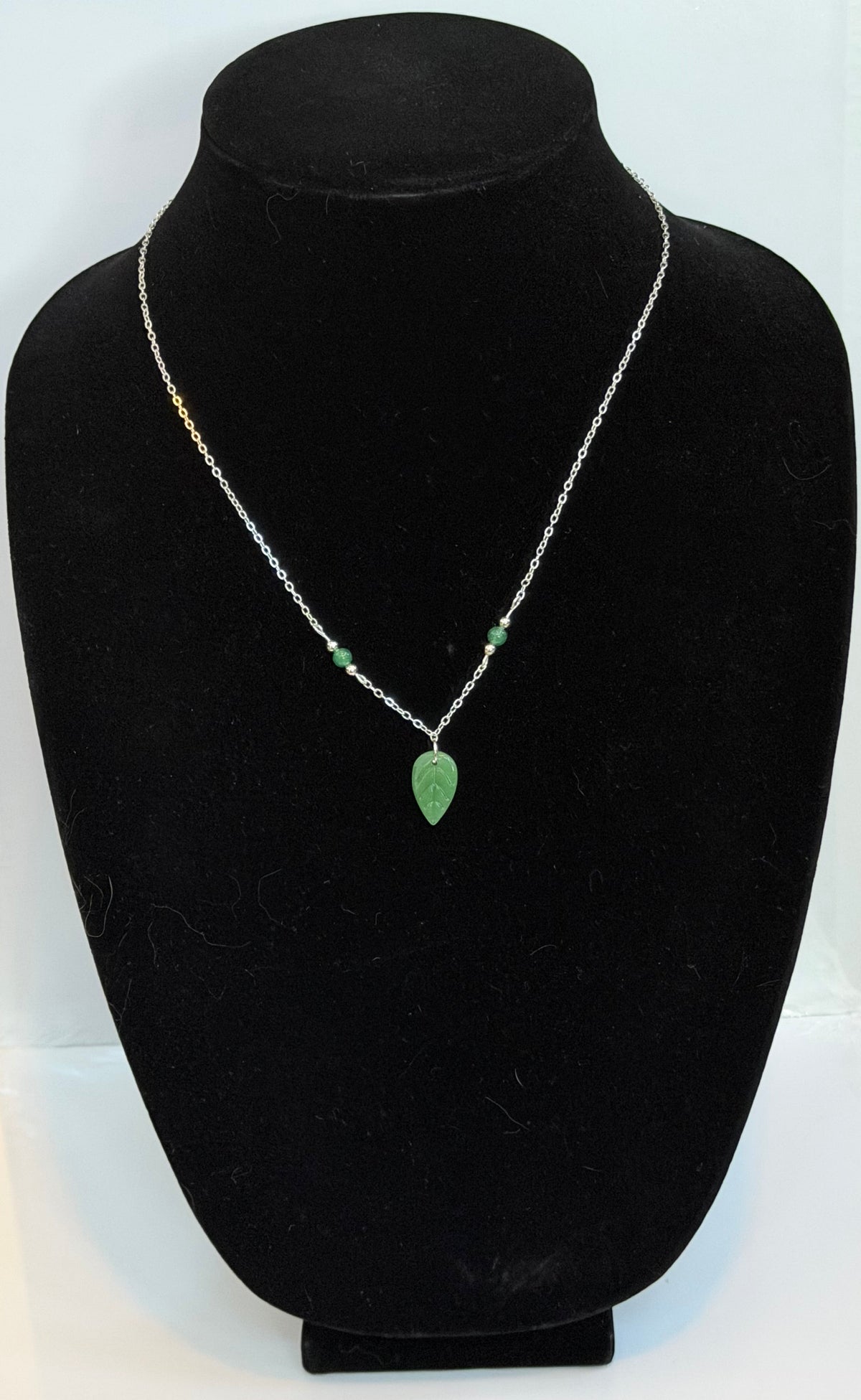 A delicate silver chain necklace with small green bead accents and a green teardrop pendant, displayed on a black velvet jewelry bust.