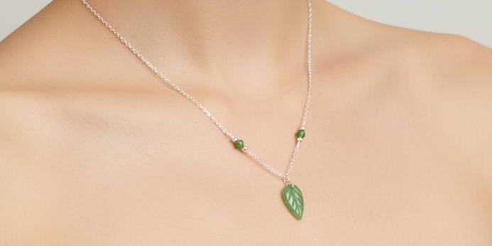 A delicate silver chain necklace worn on a model's neck, featuring a green leaf-shaped pendant and two small green bead accents.
