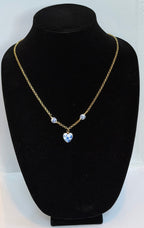 A gold chain necklace with a heart-shaped blue pendant and two small round blue stones, displayed on a black velvet jewelry bust against a white backing board.