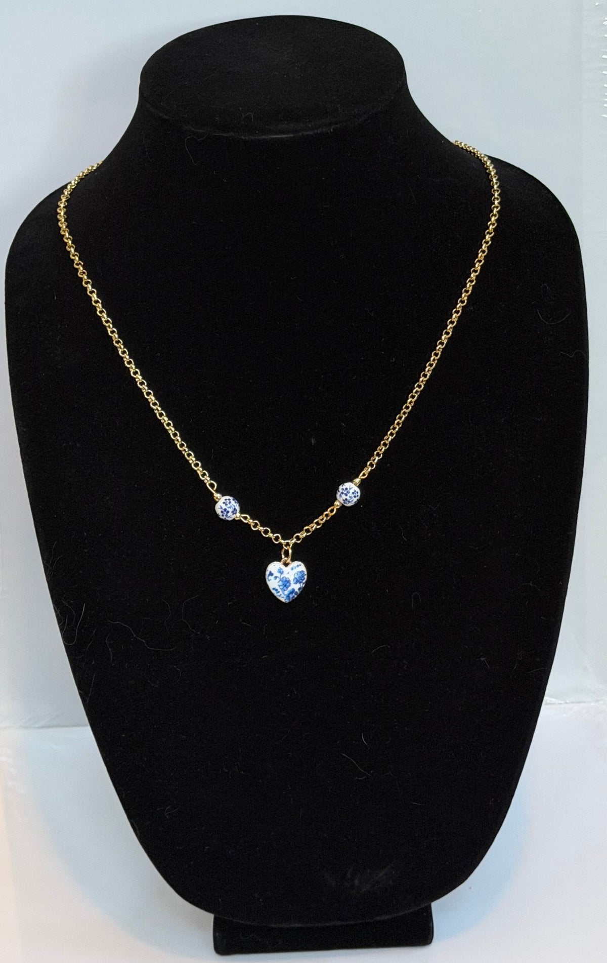A gold chain necklace with a heart-shaped blue pendant and two small round blue stones, displayed on a black velvet jewelry bust against a white backing board.