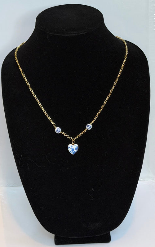 A gold chain necklace with a heart-shaped blue pendant and two small round blue stones, displayed on a black velvet jewelry bust against a white backing board.