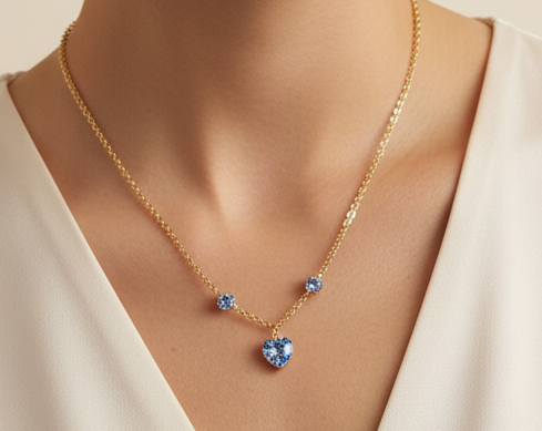 A gold chain necklace with a heart-shaped blue crystal pendant and two smaller blue crystal accents, shown worn on a model's neck.