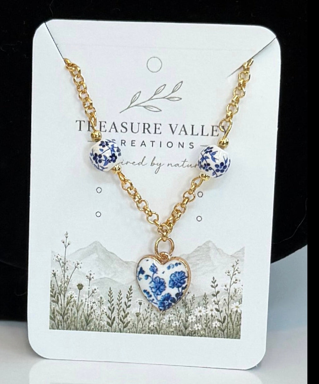 Treasure Valley Creations necklace featuring a gold chain with three blue and white floral bead-like charms and a matching heart-shaped pendant, displayed on a branded white card.
