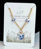 Treasure Valley Creations necklace featuring a gold chain with three blue and white floral bead-like charms and a matching heart-shaped pendant, displayed on a branded white card.