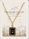 A gold chain necklace with a rectangular black pendant featuring pink flowers and green leaves, displayed on a cream-colored card labeled 'Treasure Valley Creations'.