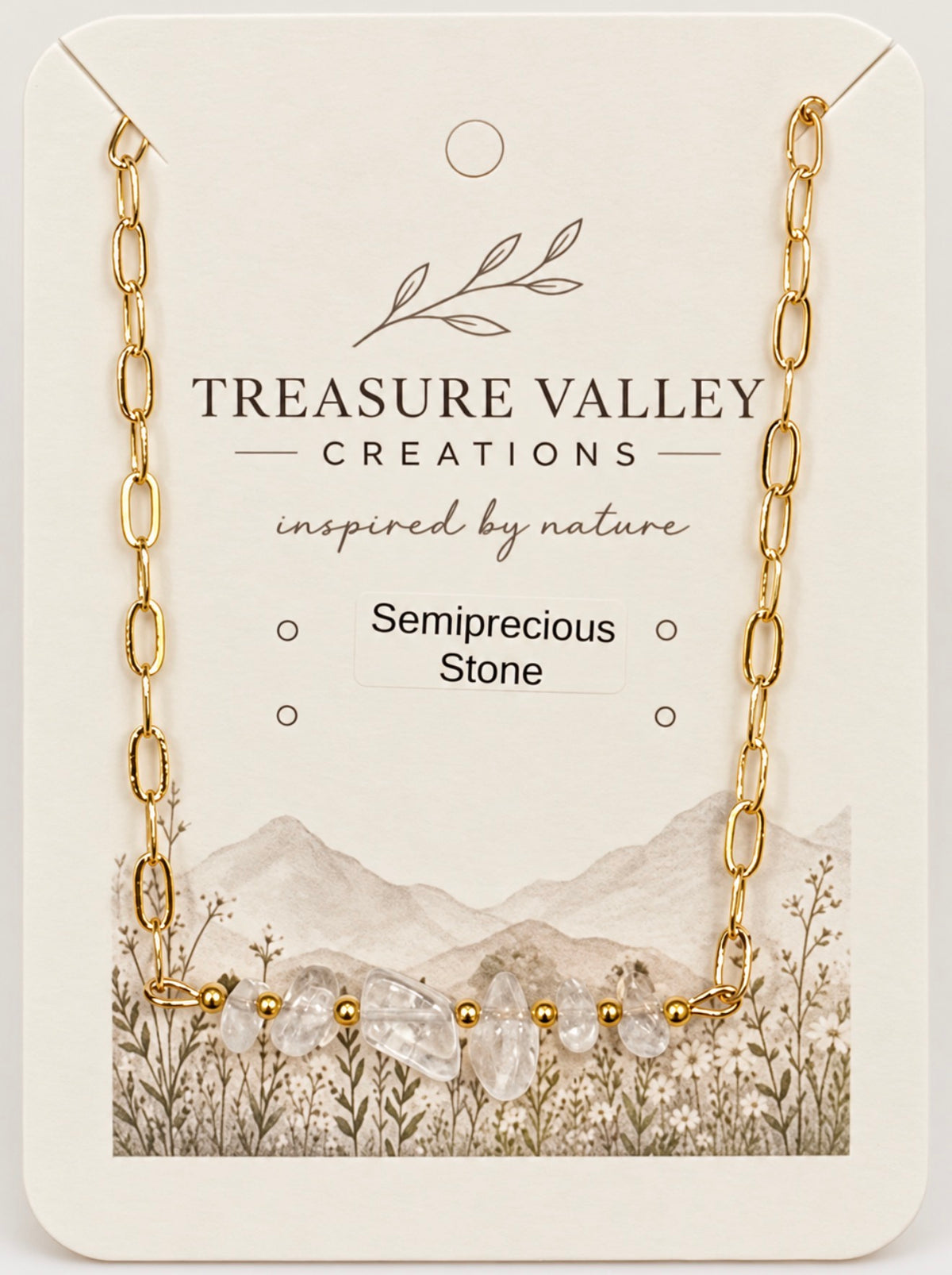 A gold chain necklace with a row of clear, faceted semiprecious stones, displayed on a cream-colored backing card. The backing card features the brand name 'Treasure Valley Creations' and a watercolor illustration of mountains and wildflowers.