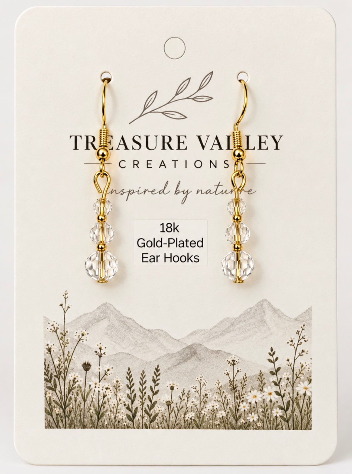 Treasure Valley Creations gold-plated ear hooks with clear faceted crystal drops, displayed on a cream-colored branded card with a mountain and wildflower illustration.