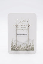 A pair of silver hypoallergenic hoop earrings with long chain links, displayed on a white backing card from the brand Treasure Valley Creations. The backing card features a botanical and mountain illustration.