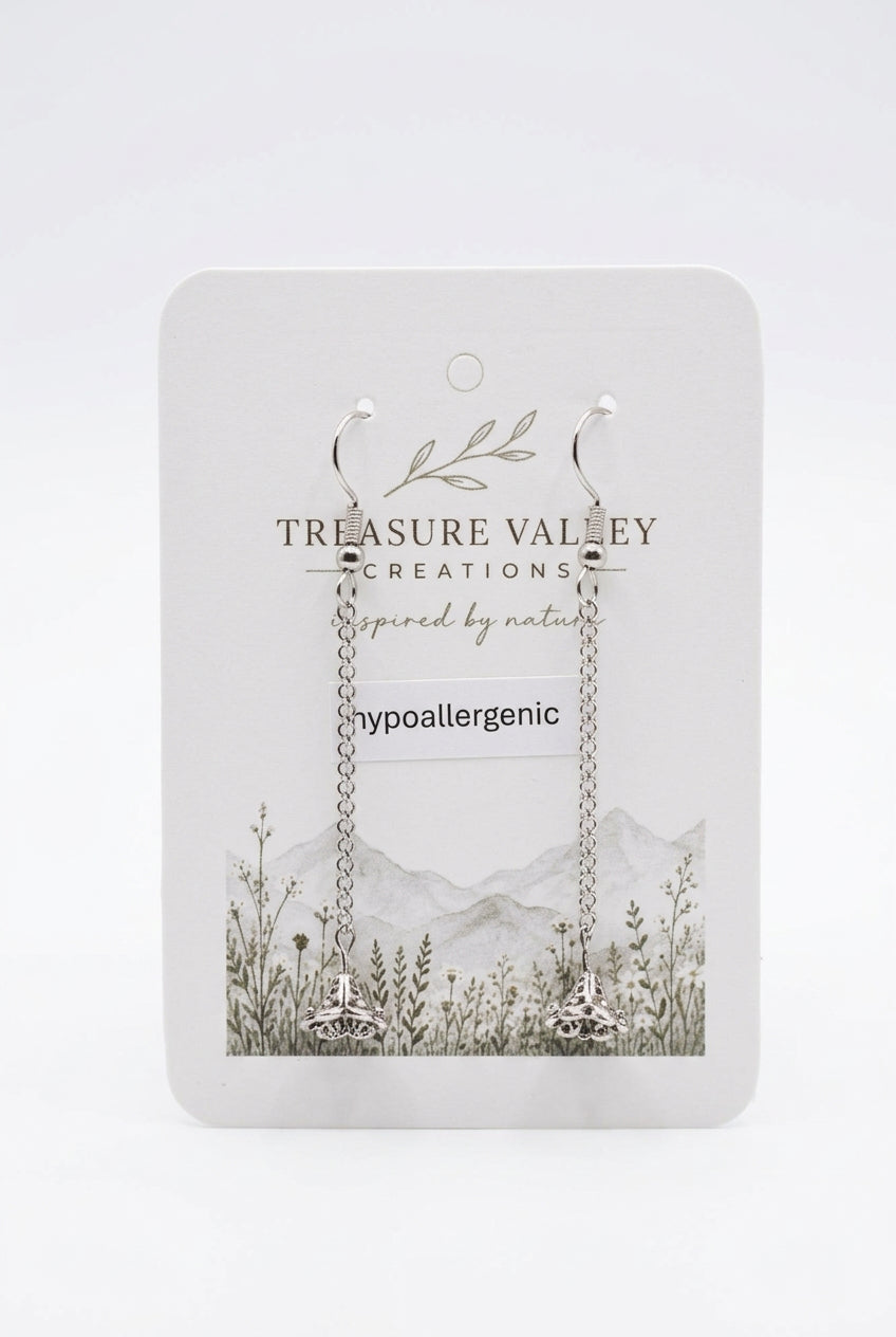 A pair of silver hypoallergenic hoop earrings with long chain links, displayed on a white backing card from the brand Treasure Valley Creations. The backing card features a botanical and mountain illustration.