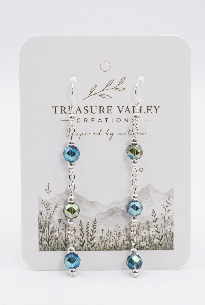 A pair of dangling earrings displayed on a white backing card. The earrings have silver-toned metal chains with faceted beads in shades of blue, teal, and olive green.