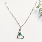 A silver chain necklace featuring a minimalist outline pendant of the state of New Hampshire, adorned with a small, faceted green gemstone, displayed on a textured white surface with a touch of green foliage in the corner.