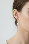 A close-up of a model's ear wearing a delicate gold dangle earring with a white pearl and a short chain.