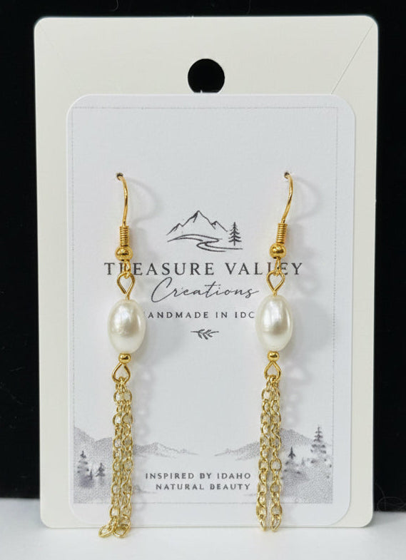 A pair of gold-tone dangle earrings with white pearl beads and chain drops, displayed on a white backing card that reads 'Treasure Valley Creations'. The card is set against a black background with a white surface below.