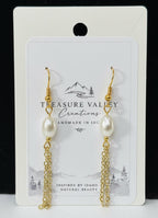 A pair of gold-tone dangle earrings with white pearl beads and chain drops, displayed on a white backing card that reads 'Treasure Valley Creations'. The card is set against a black background with a white surface below.