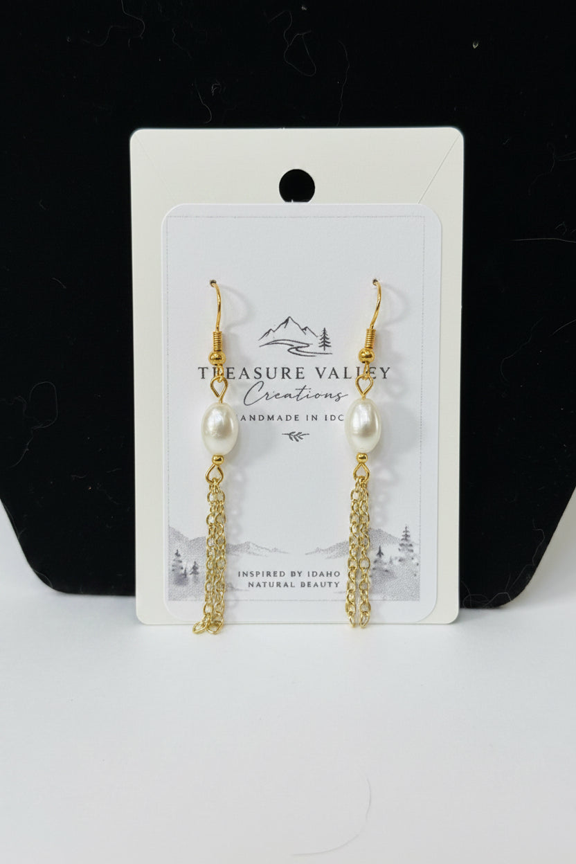 A pair of gold-tone dangle earrings with white pearl beads and chain drops, displayed on a white backing card that reads 'Treasure Valley Creations'. The card is set against a black background with a white surface below.