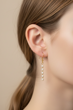 A close-up of a model's ear wearing a delicate gold dangle earring with a vertical line of small white pearls against a soft beige background.