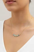 A close-up of a model wearing a delicate gold chain necklace with a curved row of small teal and white beads at the center, paired with a white V-neck top.