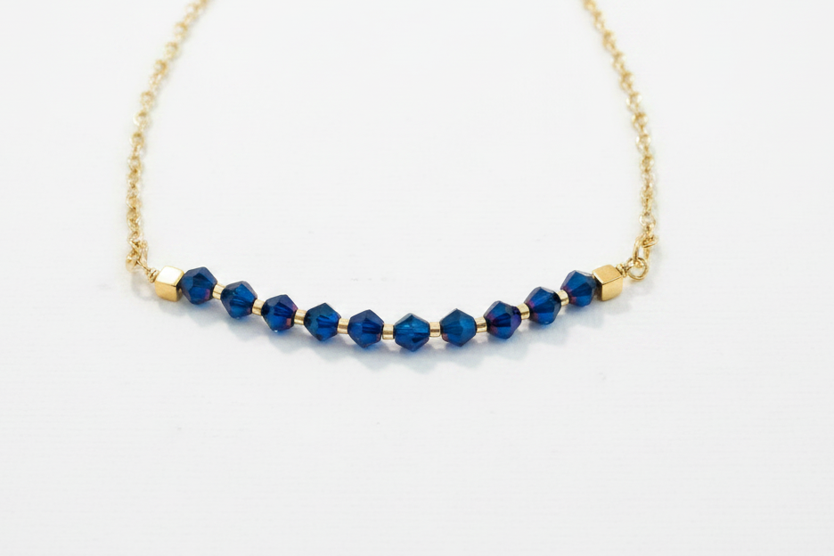 A delicate gold chain necklace featuring a curved row of faceted dark blue beads separated by small gold spacer beads, displayed on a clean white background.