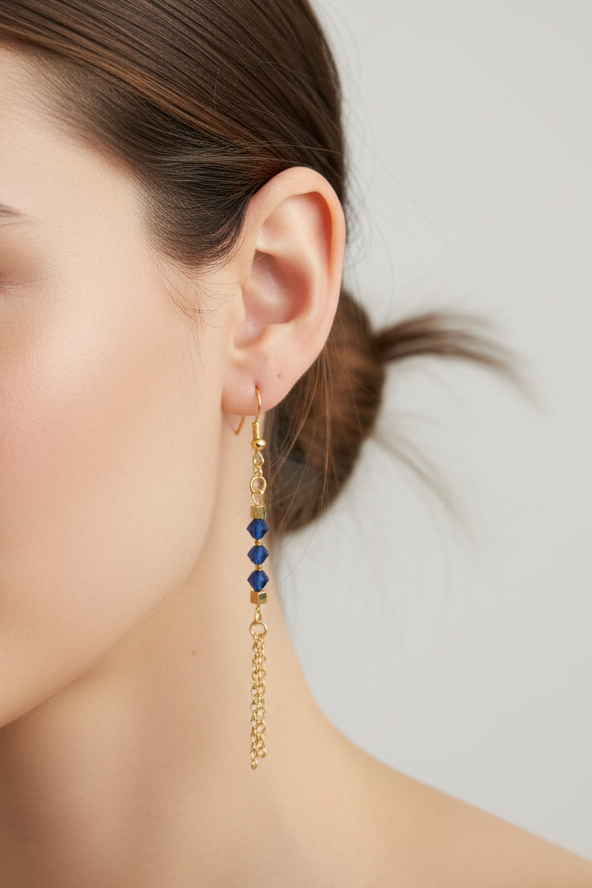 A close-up of a model's ear wearing a long gold dangle earring with three small blue faceted beads and a dangling chain.