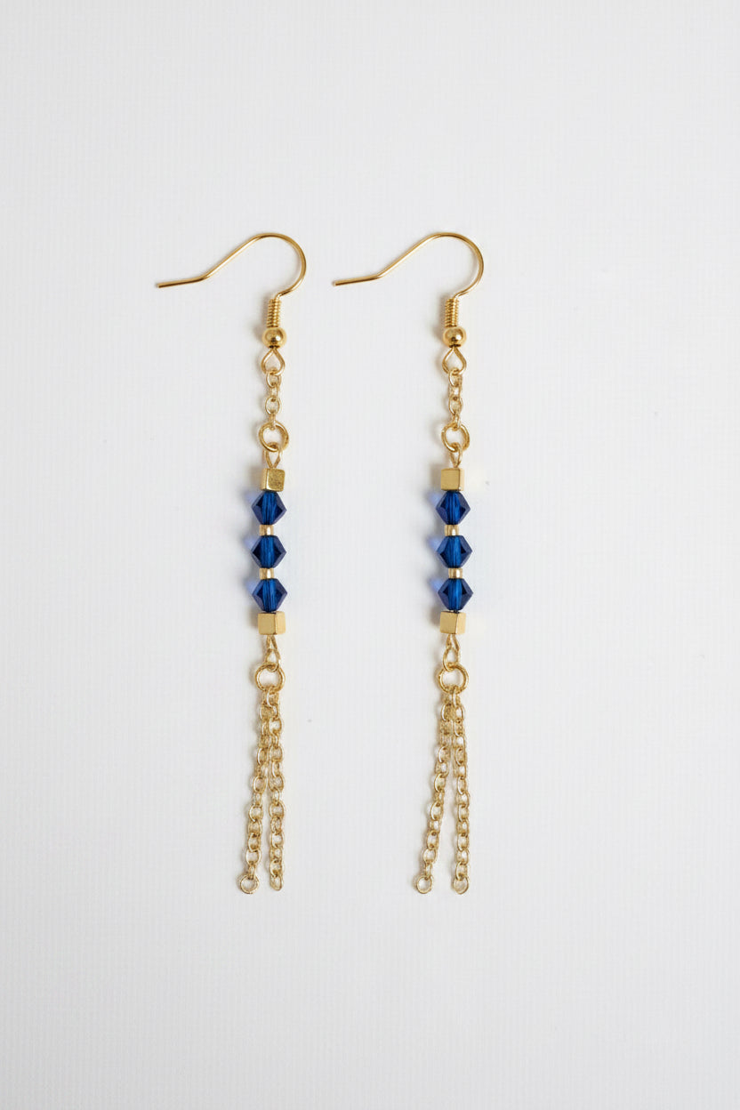 A pair of gold dangle earrings with three faceted dark blue beads on each earring, hanging from gold fishhook wires and short chain drops, shown against a plain light gray background.