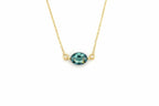 A delicate gold chain necklace featuring an oval-cut teal gemstone pendant with small gold loops on either side, shown against a plain white background.
