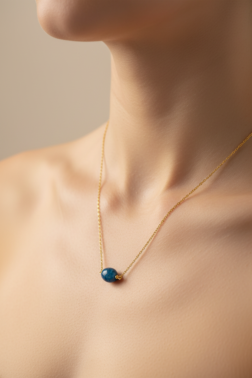 A delicate gold chain necklace with a small blue bead pendant, shown worn on a model's neck against a soft beige background.