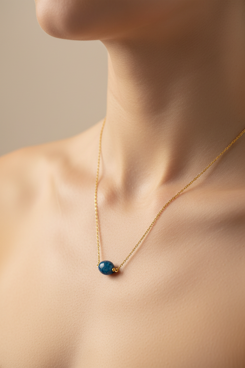 A delicate gold chain necklace with a small blue bead pendant, shown worn on a model's neck against a soft beige background.