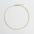 A delicate gold chain necklace with a small bead-like link pattern, a lobster clasp, and an extender chain, laid flat on a neutral light gray background.