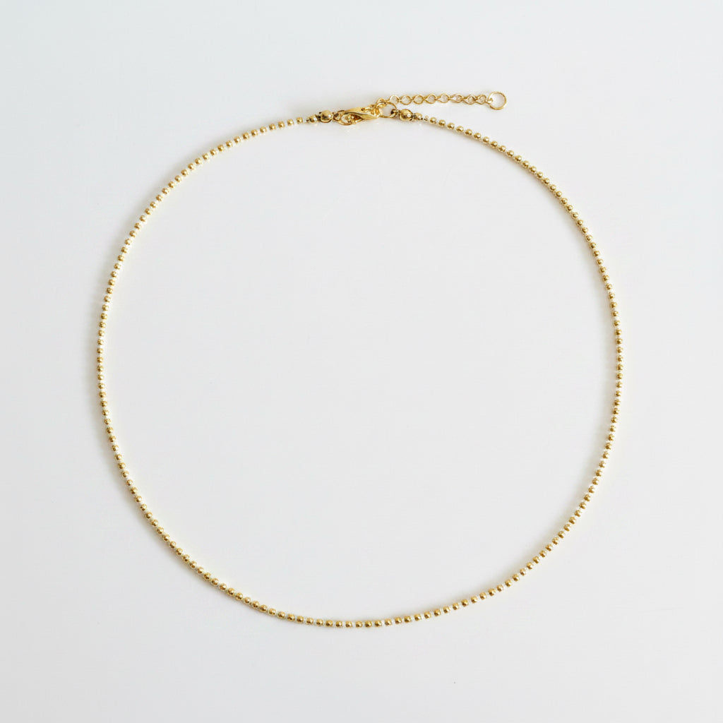 A delicate gold chain necklace with a small bead-like link pattern, a lobster clasp, and an extender chain, laid flat on a neutral light gray background.