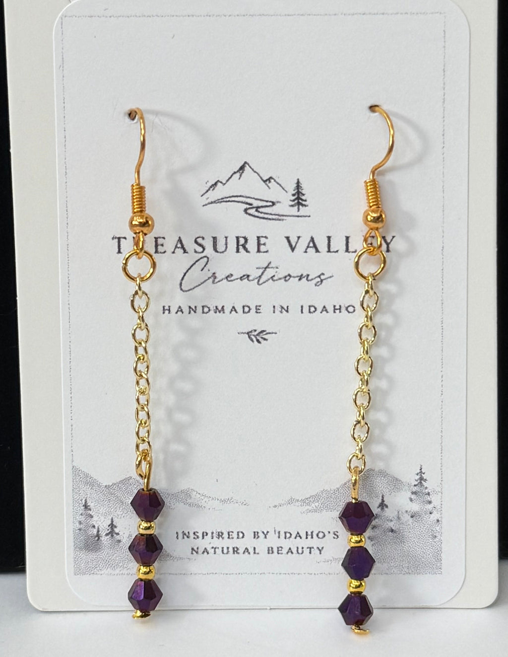 A pair of gold dangle earrings with small purple beads, displayed on a white card labeled Treasure Valley Gemstones, set against a black and white background.