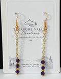 A pair of gold dangle earrings with small purple beads, displayed on a white card labeled Treasure Valley Gemstones, set against a black and white background.