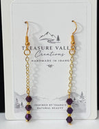 A pair of gold dangle earrings with small purple beads, displayed on a white card labeled Treasure Valley Gemstones, set against a black and white background.