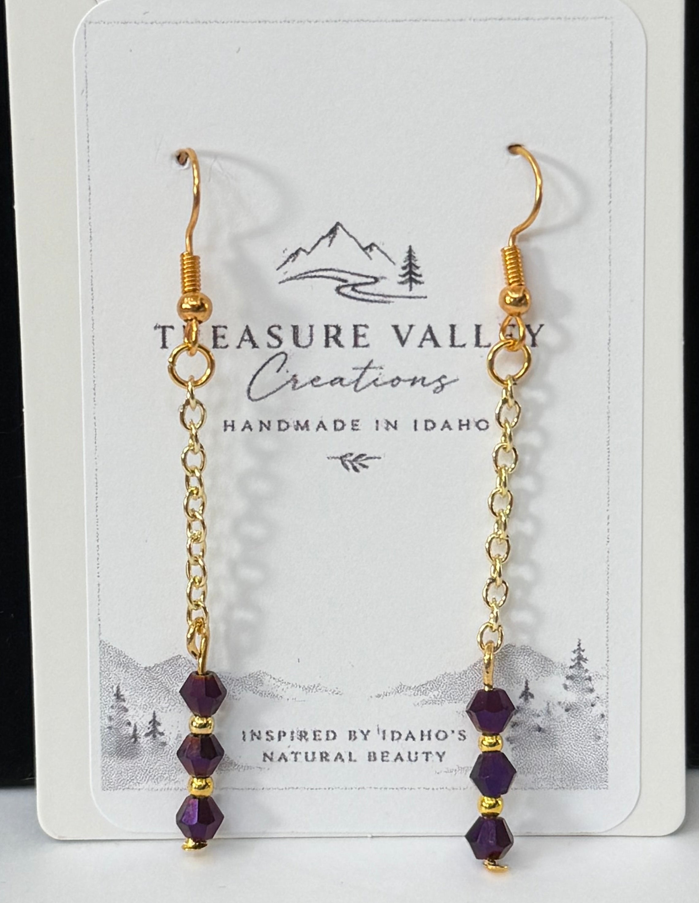 A pair of gold dangle earrings with small purple beads, displayed on a white card labeled Treasure Valley Gemstones, set against a black and white background.