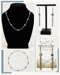 A jewelry set displayed in a collage: a silver beaded necklace on a black mannequin bust, matching drop earrings on a black stand, and the necklace and earrings on a branded card. The jewelry features iridescent blue, green, and yellow beads with silver chain links.