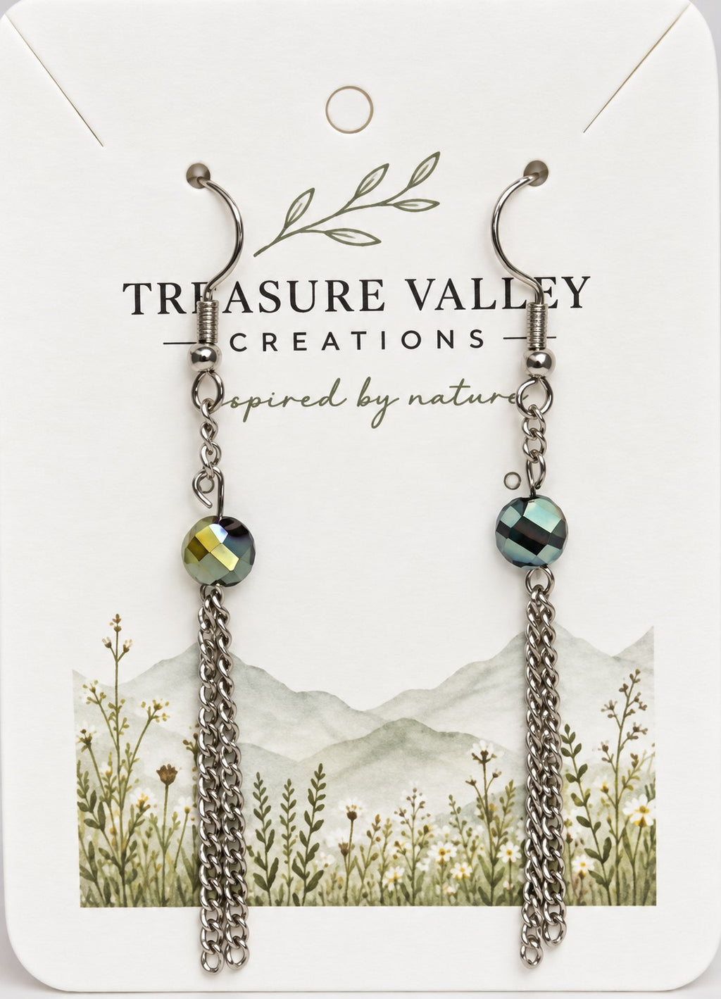 Treasure Valley Creations dangle earrings with faceted green iridescent beads and silver chain drops, displayed on a white branded card with a floral mountain illustration.