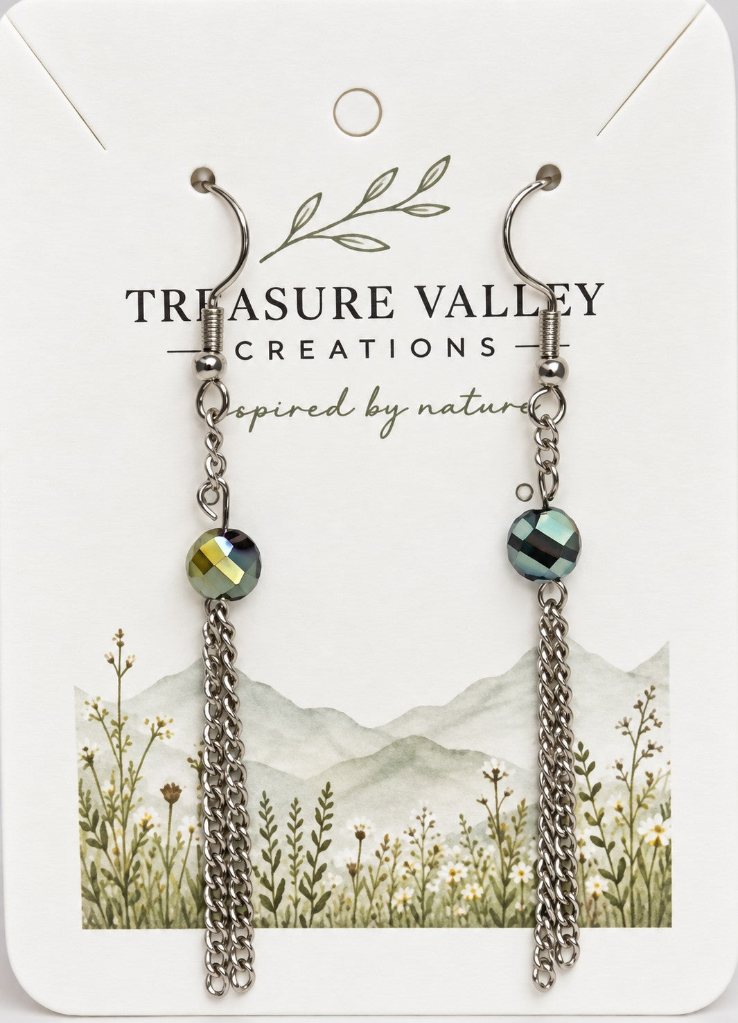 Treasure Valley Creations dangle earrings with faceted green iridescent beads and silver chain drops, displayed on a white branded card with a floral mountain illustration.