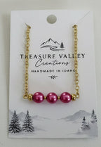 A gold chain necklace with three pinkish-red beads, displayed on a white card labeled 'Treasure Valley Creations' against a light gray background.