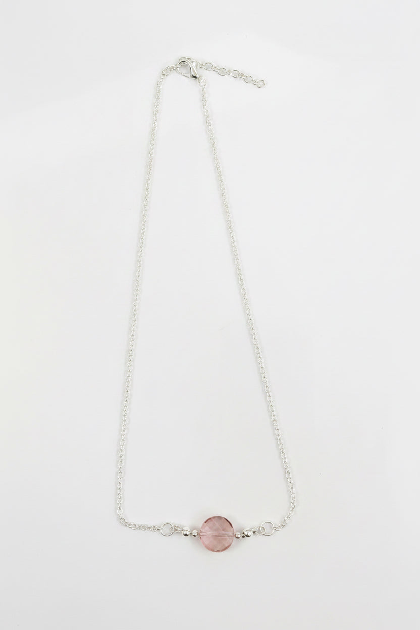 A delicate silver chain necklace featuring a single round pink bead pendant, displayed on a plain light gray background.