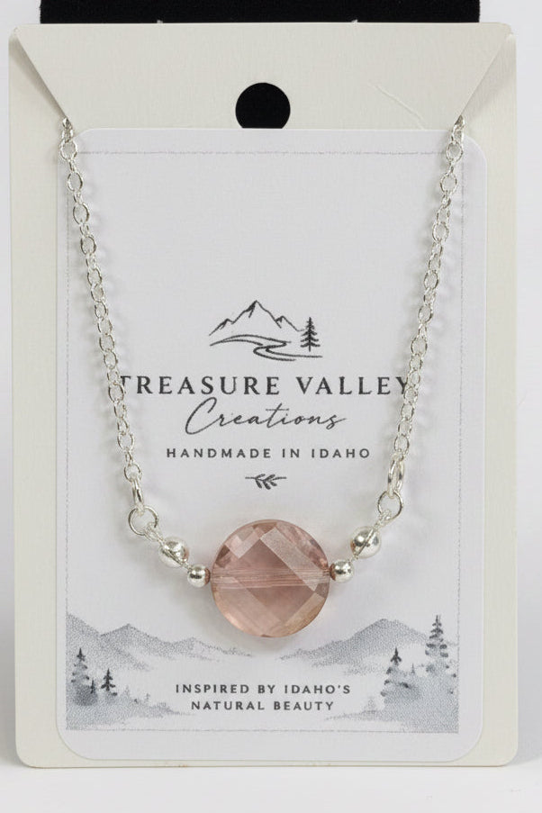 A silver chain necklace with a faceted blush pink gemstone pendant, displayed on its white backing card. The card features the brand name 'Treasure Valley Creations' and a scenic Idaho graphic.